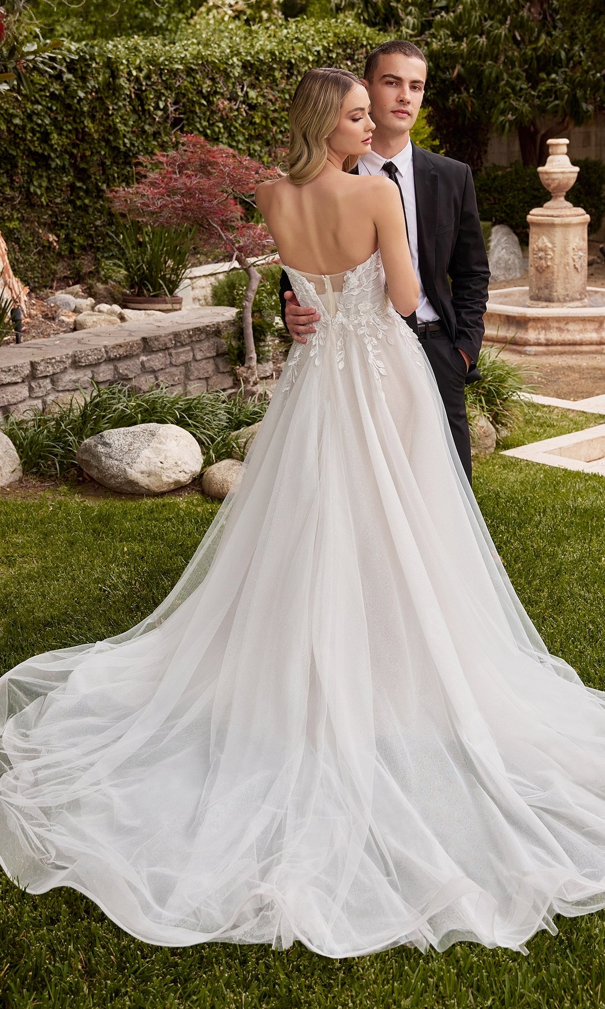 A bride wearing the Ladivine White Wedding Dress CD859W with delicate lace embroidery stands beside a suited groom in a garden, surrounded by lush greenery and elegant stonework.